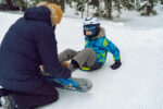 father helping little boy sitting on snow putting his feet in snowboard bindings adjusting straps.